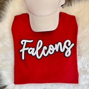 May include: A bright red t-shirt featuring the word Falcons in a white script font with a black outline. A plain white baseball cap is positioned at the top of the shirt. The design is centered on the chest of the shirt, which is made of a solid red fabric.