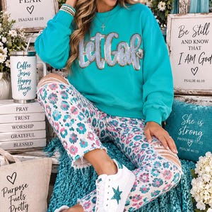 May include: A bright turquoise long-sleeve sweatshirt featuring the text Let Go in silver sequins across the chest. The outfit includes white distressed jeans with a colorful leopard print pattern and white sneakers with a glittery teal star.