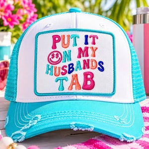 May include: A bright turquoise and white trucker hat featuring a rectangular patch with the embroidered text "PUT IT ON MY HUSBANDS TAB" in colorful, wavy letters, including a pink smiley face with dollar sign eyes on the front panel.