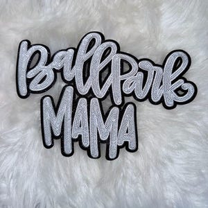 May include: A black and white decorative sign with the words "Ballpark MAMA" in a cursive font. The sign has a black outline and a white interior with a dotted pattern. The background is a fluffy white surface.