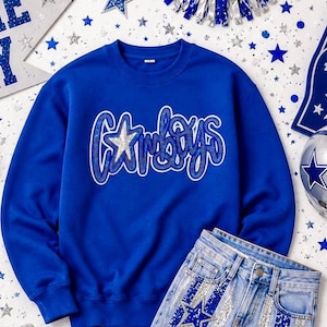 May include: A royal blue crewneck sweatshirt featuring the word Cowboys in a white-outlined, blue sequined script font with a silver sequined star replacing the letter C. The long-sleeved pullover has a ribbed collar, cuffs, and hemline.