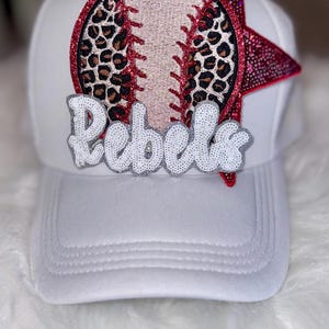 May include: White baseball cap with a baseball design and the word "Rebels". The baseball design features a leopard print pattern and red glitter accents. The word "Rebels" is in white sequins.