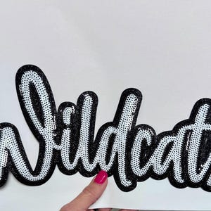 May include: A sequined patch with the word "Wildcats" in a script font. The letters are white with a black outline, and the sequins add a shimmering effect. The patch is likely for apparel or accessories.