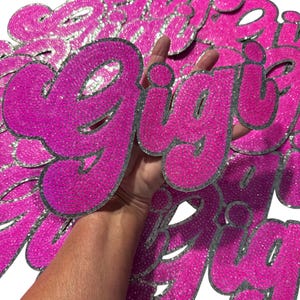 Pode incluir: Um monte de patches de ferro de brilho rosa e prata que soletram a palavra "girl".