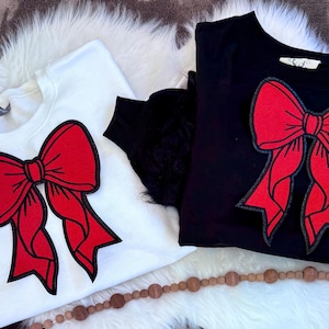 Puede incluir: Dos sudaderas, una blanca y otra negra, cada una con un diseño de lazo rojo grande. Los lazos tienen un contorno negro. Las sudaderas se exhiben sobre una superficie blanca y esponjosa, con una guirnalda de cuentas de madera.