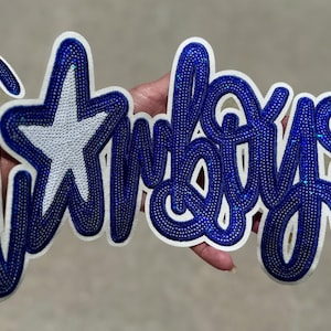 May include: A white and blue Cowboys logo with a star and script lettering. The word "Cowboys" is written in a cursive font with blue rhinestones. The star is white with a blue outline. The logo is held up by a hand.