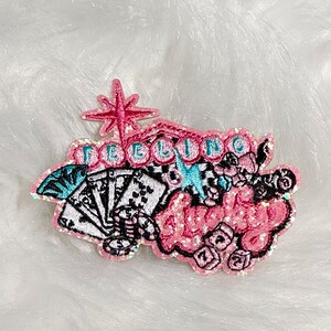 May include: Embroidered patch with the words "Feeling Lucky" in pink glitter lettering. The patch features playing cards, dice, and a starburst design, with accents of black, white, and teal. The patch is on a white, fluffy surface.