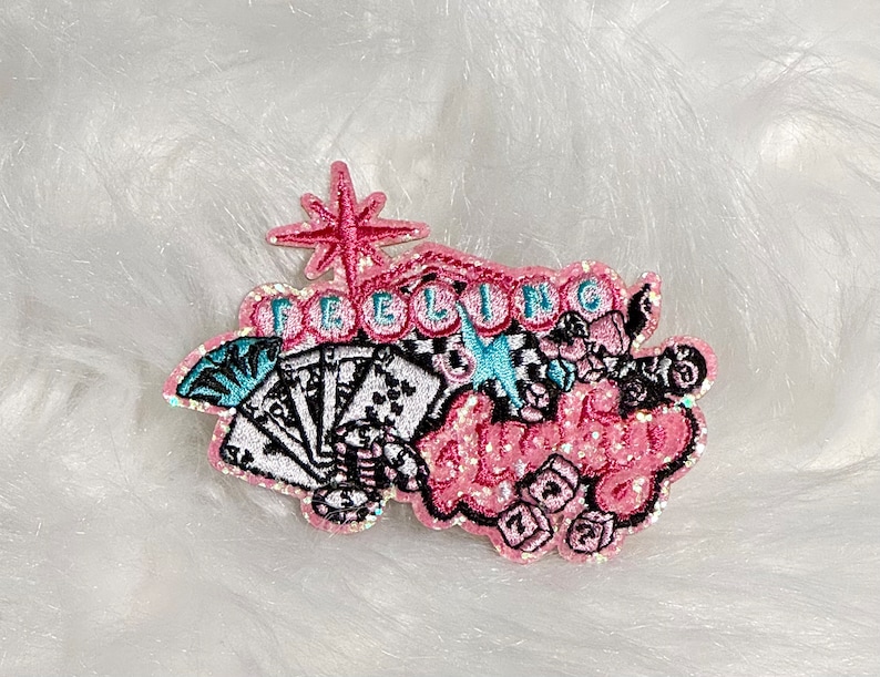 May include: Embroidered patch with the words "Feeling Lucky" in pink glitter lettering. The patch features playing cards, dice, and a starburst design, with accents of black, white, and teal. The patch is on a white, fluffy surface.