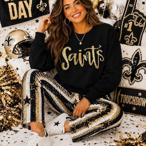 May include: A black long-sleeve sweatshirt featuring the word Saints in gold glitter script lettering, paired with trousers decorated in vertical stripes of black, white, and gold sequins with black star appliques and white sneakers.
