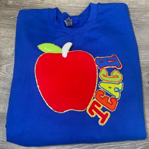 Könnte beinhalten: Ein königsblauer Pullover mit einem roten Filzapfel und dem Wort "TEACH" in Regenbogen-Glitzerbuchstaben.