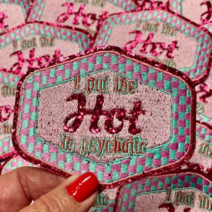 May include: Pink and green checkered iron-on patch with pink glitter lettering that reads "I put the hot in psychotic".