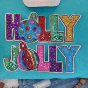 Puede incluir: Una sudadera turquesa con las palabras "HOLLY JOLLY" en letras con lentejuelas. Las letras están delineadas en plata y presentan diseños de adornos navideños con lentejuelas de colores. Una gorra de béisbol blanca es visible en la parte superior.