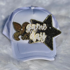 May include: White trucker hat with a mesh back and a curved brim. The hat features a gold fleur-de-lis, a black star, and the words "Game Day" in gold and white sequins. A stylish accessory for game day.