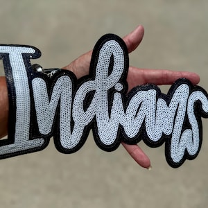 May include: A black and white sequined patch in a script font that spells "Indians". The patch has a black border and is held by a hand. The sequins are white and create a shimmering effect.