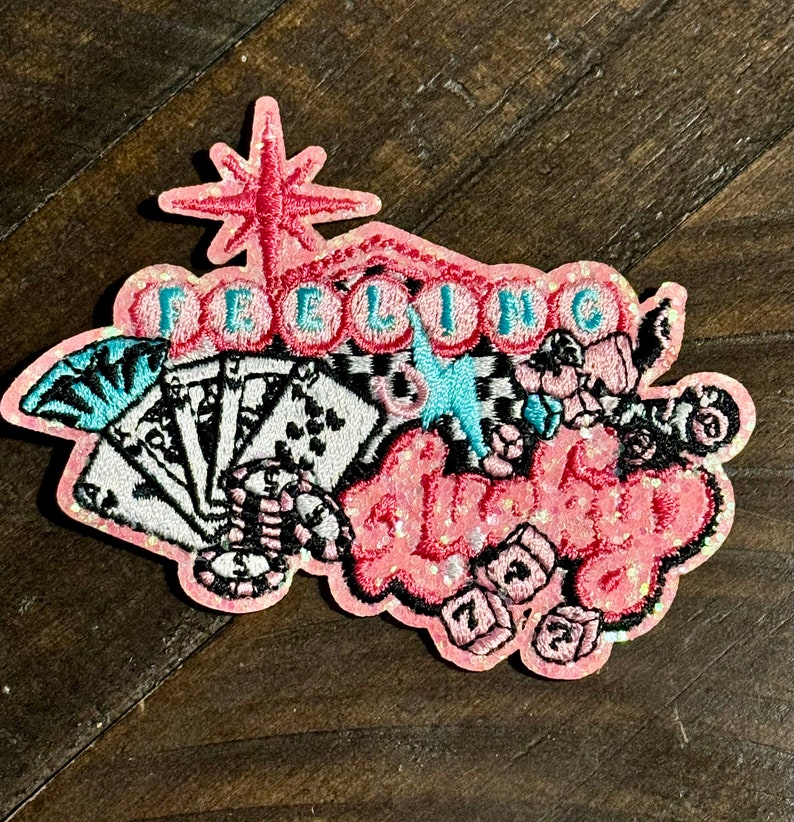 May include: A pink embroidered patch with the words "Feeling Lucky" in a cursive font. The patch features playing cards, dice, and a starburst design. The patch is embellished with glitter.