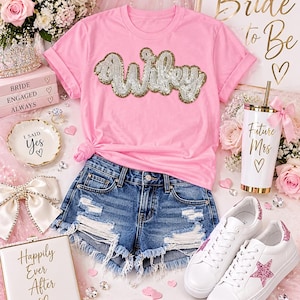 May include: A light pink t-shirt with the word Wifey in gold and silver sequins, paired with distressed blue denim shorts, white sneakers with pink glitter stars, a white tumbler with Future Mrs text, and a white clutch with Happily Ever After text.