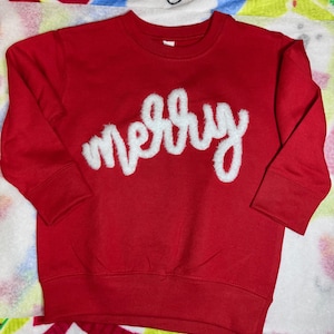 May include: A red sweatshirt with the word "merry" in white, fluffy lettering across the chest. The long-sleeved top is made of a soft material and is suitable for children.