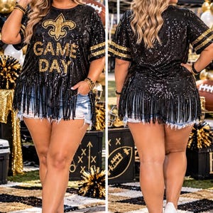 May include: A black sequined short-sleeve shirt featuring a gold fleur-de-lis emblem above the text GAME DAY in gold lettering. The shirt has gold striped sleeve cuffs and a long black fringe hemline, paired with distressed blue denim shorts.