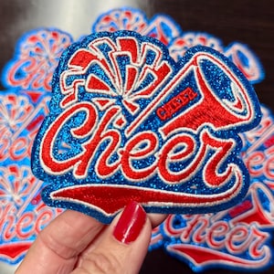 Peut inclure: Écussons brodés bleus, rouges et blancs avec le mot "Cheer" en écriture cursive. Les écussons présentent un mégaphone et un motif d'éclatement, avec des détails pailletés. Plusieurs écussons similaires sont visibles.