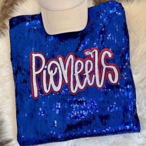 May include: A bright blue sequinned sweatshirt featuring the word Pioneers in white script with a red outline, paired with a plain white baseball cap. The garment is covered in shimmering blue sequins that create a textured, reflective surface.
