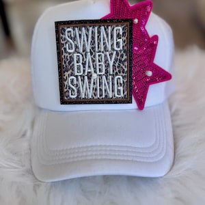 Puede incluir: Gorra de camionero blanca con un parche con estampado de leopardo que dice "SWING BABY SWING" en letras bordadas. Dos estrellas rosas con lentejuelas están unidas al costado del sombrero. La gorra tiene una visera curva y una parte trasera de malla.