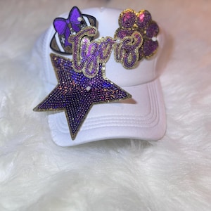 Op de afbeelding: Witte baseballcap versierd met een paarse ster, een boog in de vorm van een voetbal en een pootafdruk, allemaal versierd met glinsterende strass-steentjes. Het woord "Tigers" is in gouden letters geschreven. Een modieus accessoire.