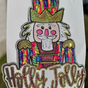 May include: A colorful, sequined Christmas-themed patch featuring a nutcracker soldier with a gold crown and the words "Holly Jolly". The nutcracker's uniform is a mix of rainbow colors, and the text is outlined in gold.