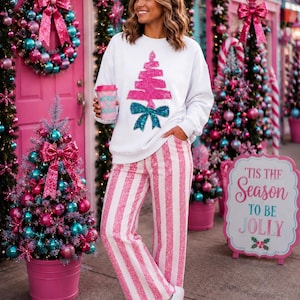 May include: A white long-sleeve sweatshirt featuring a pink sequined Christmas tree graphic with a teal bow, paired with pink and white vertical striped trousers and white trainers with pink accents, while holding a pink cup labelled Merry & Bright.
