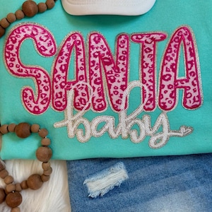 May include: A turquoise sweatshirt with the words "SANTA BABY" in pink leopard print and silver glitter trim. A white baseball cap and denim shorts are also visible. Wooden beads and a white furry surface are in the background.