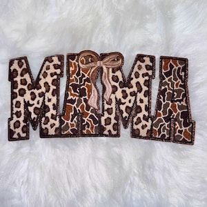 May include: Embroidered patch spelling "MAMA" in leopard print with a brown border. A bow is between the two "M" letters. The patch is on a white, fluffy surface.