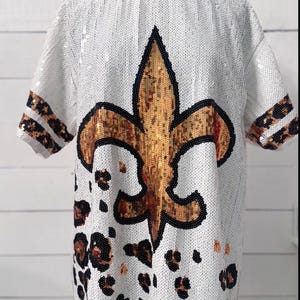 Könnte beinhalten: Weißes Pailletten-T-Shirt mit einem großen goldenen Fleur-de-Lis-Design, das schwarz umrandet ist. Die Ärmel haben ein Leopardenmuster, und der untere Teil des Shirts hat Leopardenflecken. Dieses Oberteil ist ein einzigartiges Kleidungsstück.