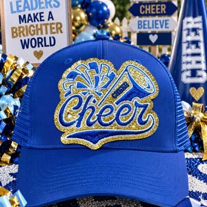 Può includere: Un cappellino da baseball blu reale con una toppa centrale ricamata con accenti glitter dorati. La toppa mostra un megafono stilizzato e un pompon sopra la parola Cheer in corsivo, con una scritta Cheer più piccola sul megafono.