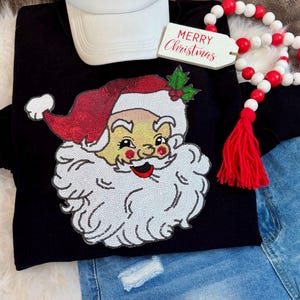 May include: Black sweatshirt featuring a sequined Santa Claus face with a red hat and white beard. A white baseball cap and a tag that says "MERRY Christmas" are also visible. Red and white beaded garland and denim shorts complete the festive ensemble.
