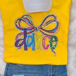 May include: A yellow sweatshirt with the word "DANCE" in colorful sequined letters. A sequined bow design is above the word. A white baseball cap is partially visible at the top. The bottom of the sweatshirt is paired with blue denim jeans.