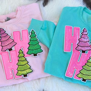 Pode incluir: Duas camisolas de gola redonda, uma rosa e outra azul-turquesa, cada uma adornada com as palavras "HO HO" em letras rosa grandes com lantejoulas. Patches de árvores de Natal em várias cores são colocados nas camisolas. As camisolas estão sobre uma superfície de pele sintética branca.