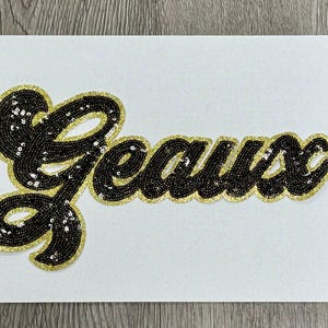 May include: A decorative patch featuring the word Geaux written in a cursive script. The letters are filled with black sequins and outlined with a thick border of shimmering gold glitter, creating a high-contrast, textured appearance.