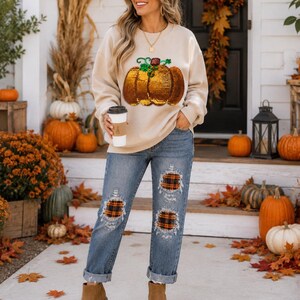 Puede incluir: Una sudadera beige de cuello redondo con un gran gráfico de calabaza de lentejuelas con hojas verdes, combinada con vaqueros azules que tienen parches desgastados con tela de cuadros naranja y negra, y botines color canela con suela gruesa.