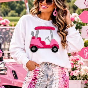 May include: A white long-sleeve sweatshirt featuring a pink golf cart graphic with a textured chenille-style finish. The sweatshirt is paired with light-wash denim jeans that have distressed knees and vertical stripes of pink and silver sequins.