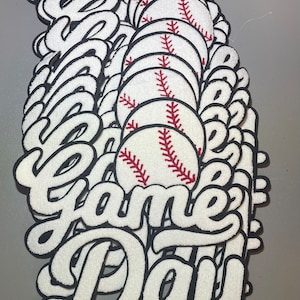 May include: A stack of white felt patches with black outlines. The patches feature the words "Game Day" in a stylized font and a red and white baseball graphic.
