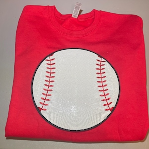 May include: Red sweatshirt with a white baseball graphic made of rhinestones. The baseball has black stitching and red laces.