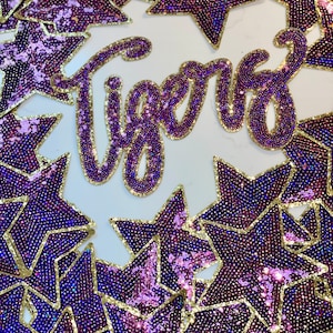 Puede incluir: Parches con forma de estrella de lentejuelas moradas y doradas con la palabra "Tigers" en el centro.