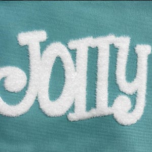 May include: Close-up of the word "JOLLY" embroidered in white, fluffy, textured letters on a teal fabric background. The letters have a playful, whimsical style, with rounded edges and a soft appearance.