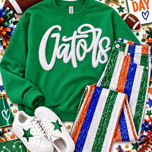 Könnte beinhalten: Ein grünes Sweatshirt mit Rundhalsausschnitt und weißem, flauschigem Schriftzug Gators, kombiniert mit einer Paillettenhose mit vertikalen Streifen in Grün, Weiß, Orange und Blau sowie weißen Sneakern mit grünen Sternen an den Seiten.