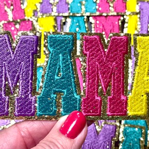 Peut inclure: Un ensemble de quatre lettres en feutre color&eacute; &eacute;pellant "MAMA" avec une bordure dor&eacute;e paillet&eacute;e. Les lettres sont brod&eacute;es et ont une texture l&eacute;g&egrave;rement en relief.