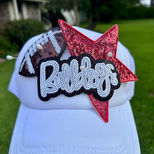 Puede incluir: Gorra de camionero blanca con parte trasera de malla, con un balón de fútbol americano, una estrella roja con lentejuelas y la palabra "Bulldogs" en un diseño en blanco y negro. La gorra tiene una visera curva.