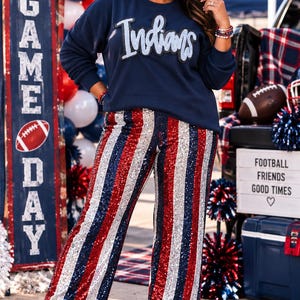 May include: A navy blue long-sleeve sweatshirt with the word Indians in white script lettering across the chest, paired with wide-leg trousers featuring vertical stripes of red, white, and blue sequins and white ankle boots.