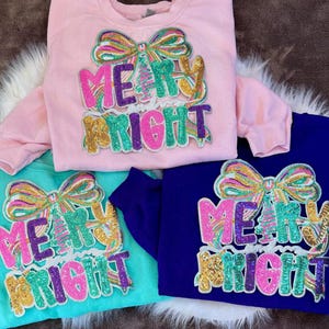 Puede incluir: Tres sudaderas en rosa, turquesa y azul marino, cada una adornada con un diseño de lentejuelas. El diseño presenta un lazo colorido y las palabras "MERRY BRIGHT" en letras de lentejuelas, perfectas para la temporada navideña.