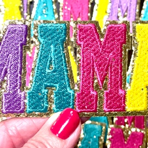 Peut inclure: Un ensemble de cinq lettres en feutre color&eacute; &eacute;pellant "MAMA" avec une bordure dor&eacute;e paillet&eacute;e. Les lettres sont violettes, turquoise, roses et jaunes.
