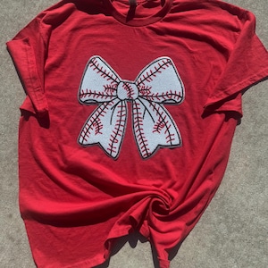 May include: Red t-shirt with a white baseball-themed bow on the front.