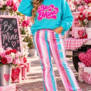 May include: A bright turquoise sweatshirt featuring the text Be Mine in pink and gold lettering, paired with high-waisted jeans covered in vertical stripes of pink, white, and turquoise sequins with distressed details and white platform sneakers.